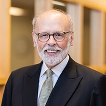 A smiling elderly man with a graying beard and glasses is dressed in a dark suit, light dress shirt, and patterned tie. He stands indoors in front of a blurred background featuring glass windows and wooden frames.