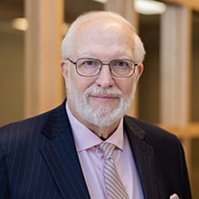 An older man with a white beard and glasses is wearing a dark pinstripe suit, a light pink shirt, and a matching pink tie. He is standing indoors in front of a blurred background with warm tones, giving a professional and approachable appearance.