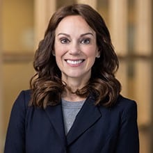 A woman with shoulder-length wavy brown hair, wearing a navy blazer and a gray top, smiles warmly at the camera. The background is softly focused with neutral-colored walls and large windows, suggesting an indoor office setting.