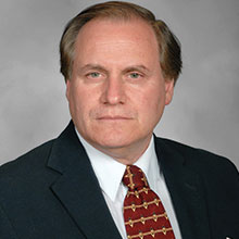 A man with short, light brown hair is wearing a dark suit, white dress shirt, and a red patterned tie. He has a serious expression and is positioned against a gray background.