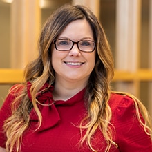 A woman with long, wavy hair stands smiling. She is wearing glasses and a red top with a decorative bow on the collar. The background is softly blurred with vertical light-colored elements, likely columns or beams.