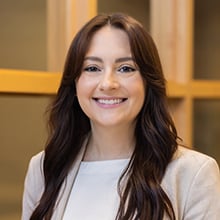 A person with long, dark hair is smiling at the camera. They are wearing a light-colored blazer over a light blouse. In the background, there are wooden window frames which suggest an indoor setting.