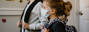 Two children wearing backpacks and face masks stand indoors. The child on the right has blonde hair tied up and wears a white mask, while the other child’s face is partially visible. They appear to be preparing for school. A door and wall are visible in the background.