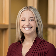 A woman with long blonde hair is smiling at the camera. She is wearing a maroon blouse, and the background features a wooden partition with a softly blurred, warm-toned backdrop.