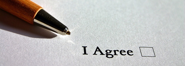 A close-up of a ballpoint pen pointing towards a checkbox next to the text "I Agree" on a piece of paper. The checkbox is empty, hinting at agreement to a noncompete clause.