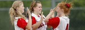 three girls in red and white softball jerseys celebrating a win