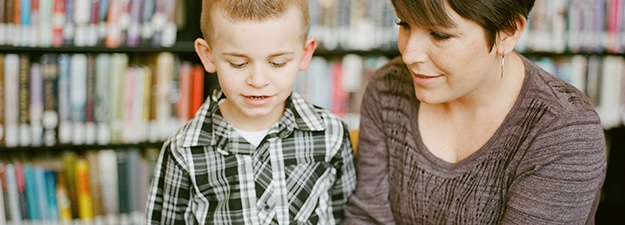 A young boy in a plaid shirt is sitting next to a woman with short dark hair. They are reading a book together in a library, with bookshelves filled with books in the background, perhaps following some teacher training guidelines for effective literacy development.