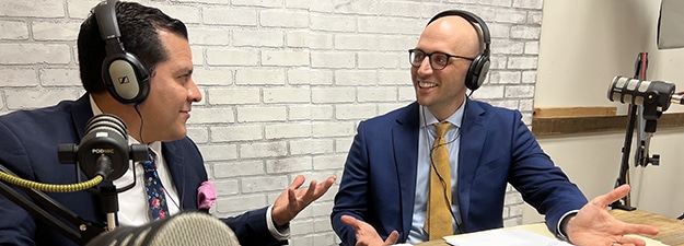 Two men in suits are sitting at a desk with microphones, wearing headphones, and engaged in a lively conversation. One man gestures with his hands while speaking, and both are smiling. They are in a room with a white brick wall backdrop.