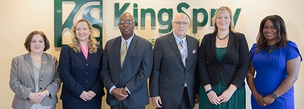 A group of six professionally dressed individuals stand side-by-side in front of a wall with the "KingSpry" logo. There are three men and three women, all smiling at the camera. The individuals are wearing business attire in a mix of suits, ties, and dresses.