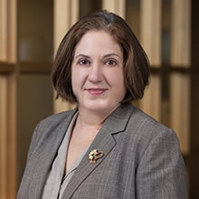 A woman with short brown hair, wearing a gray blazer with a decorative pin, looks at the camera. She stands indoors in front of a wooden paneled background.