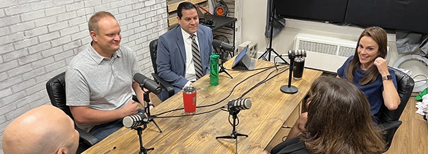 Five people sitting around a wooden table with microphones, engaged in a discussion. The room has a white brick wall background, and various items are visible on shelves. Two women and three men appear to be participating in a podcast or meeting.