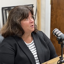 A woman with shoulder-length brown hair, wearing a black blazer over a white and black striped shirt, is sitting beside a microphone. She appears to be speaking or engaged in a conversation in a recording studio or podcast setting.