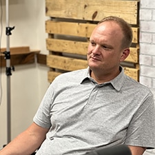 A person with short hair and a gray polo shirt sits indoors. The background has wooden pallets and a white brick wall. The individual appears to be in a casual setting, possibly a studio or workspace.