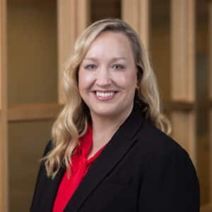 A smiling woman with long blonde hair wearing a black blazer and red blouse stands in front of a wooden background.