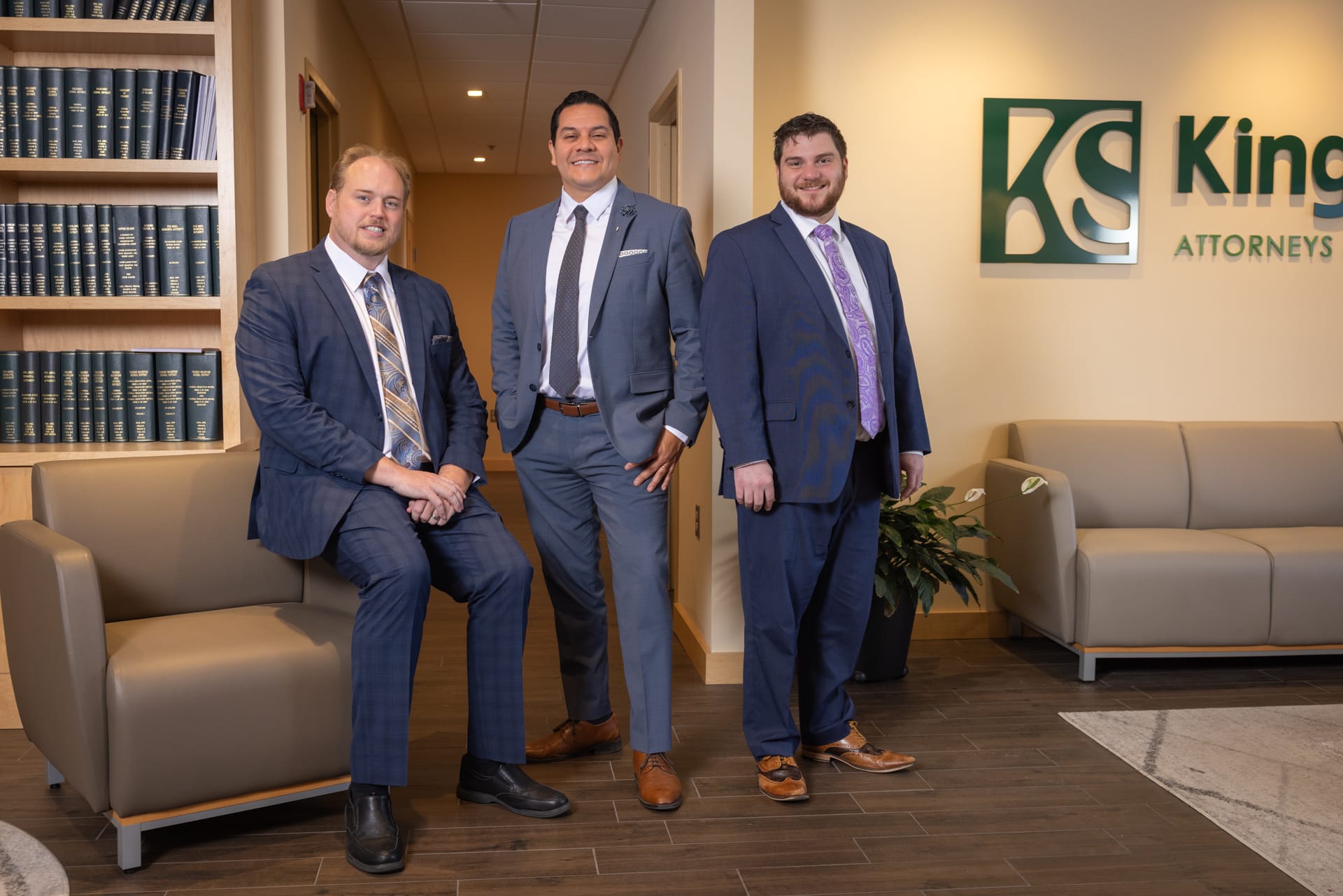 Three men in suits posed in an office lobby with a law firm's signage on the wall. Two are standing, and one is sitting on a couch. The room features bookshelves and seating, creating a professional atmosphere.